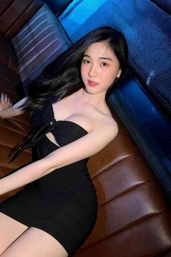 Beautiful Asian woman in black dress reclining on a brown leather couch, looking at the camera.