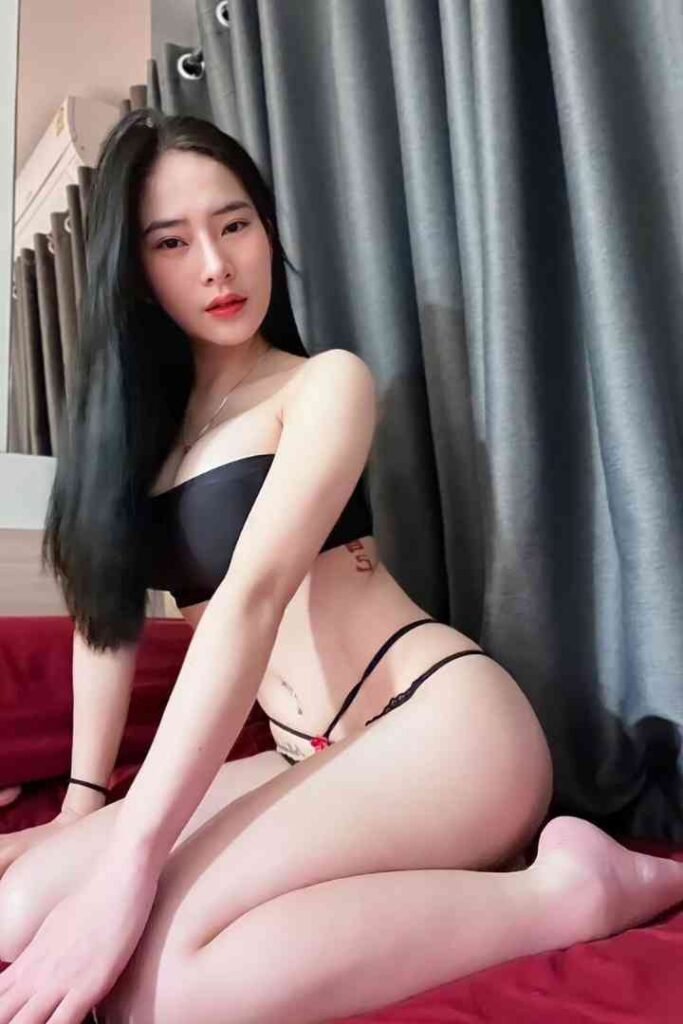 Attractive Asian woman in black strapless top and thong kneeling on bed in bedroom setting.