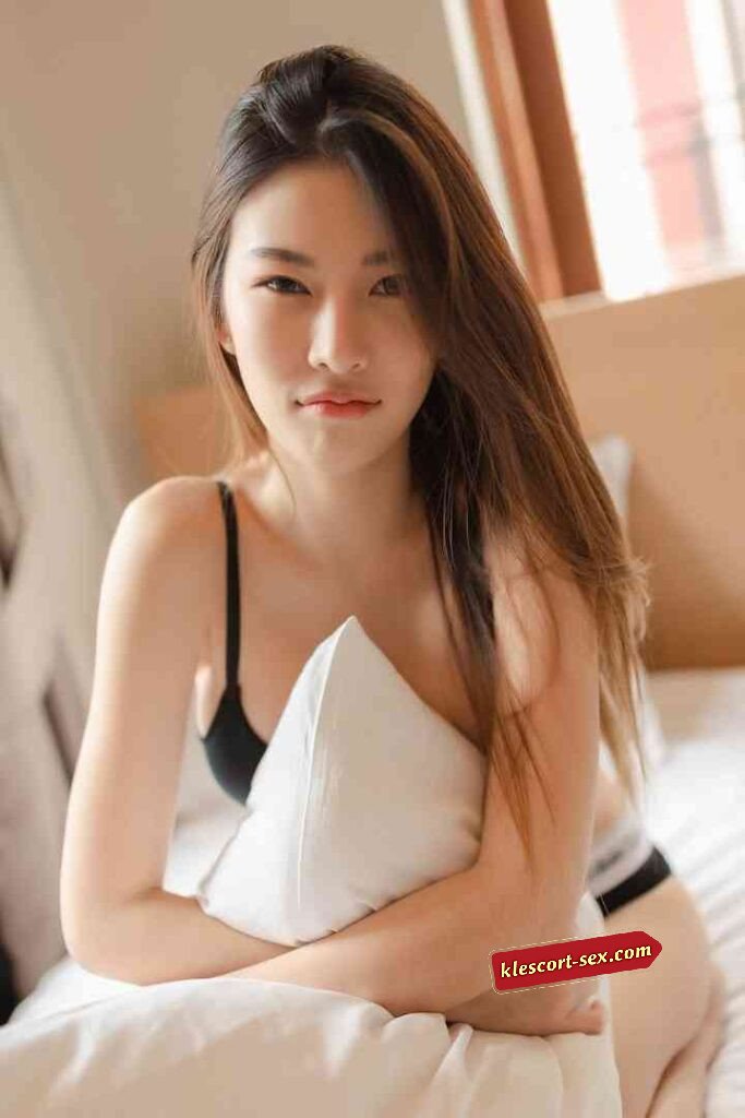 Asian woman in bra holding pillow, looking at camera. Bedroom setting, warm lighting.