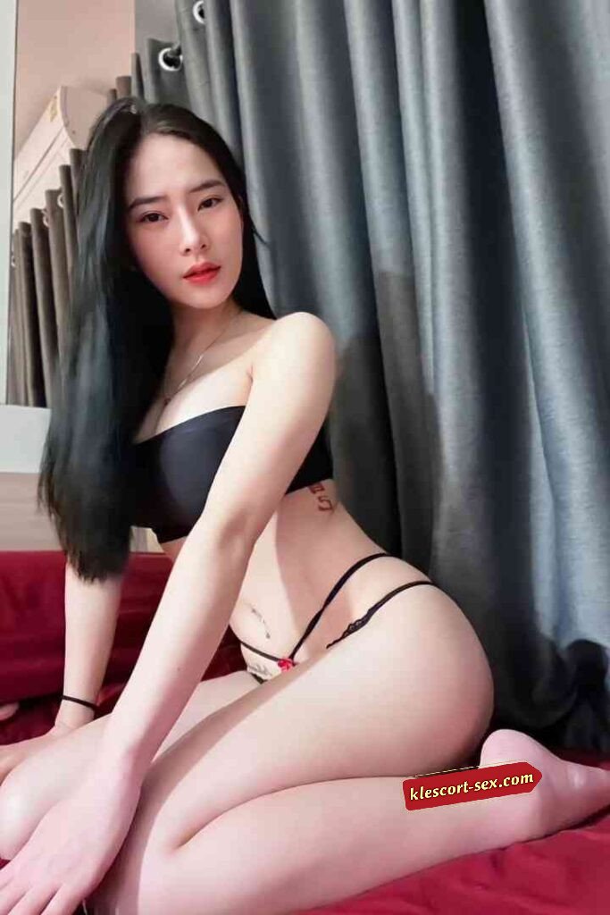 Attractive Asian woman in lingerie sitting on a red bed, posing for the camera. Black hair and fair skin.
