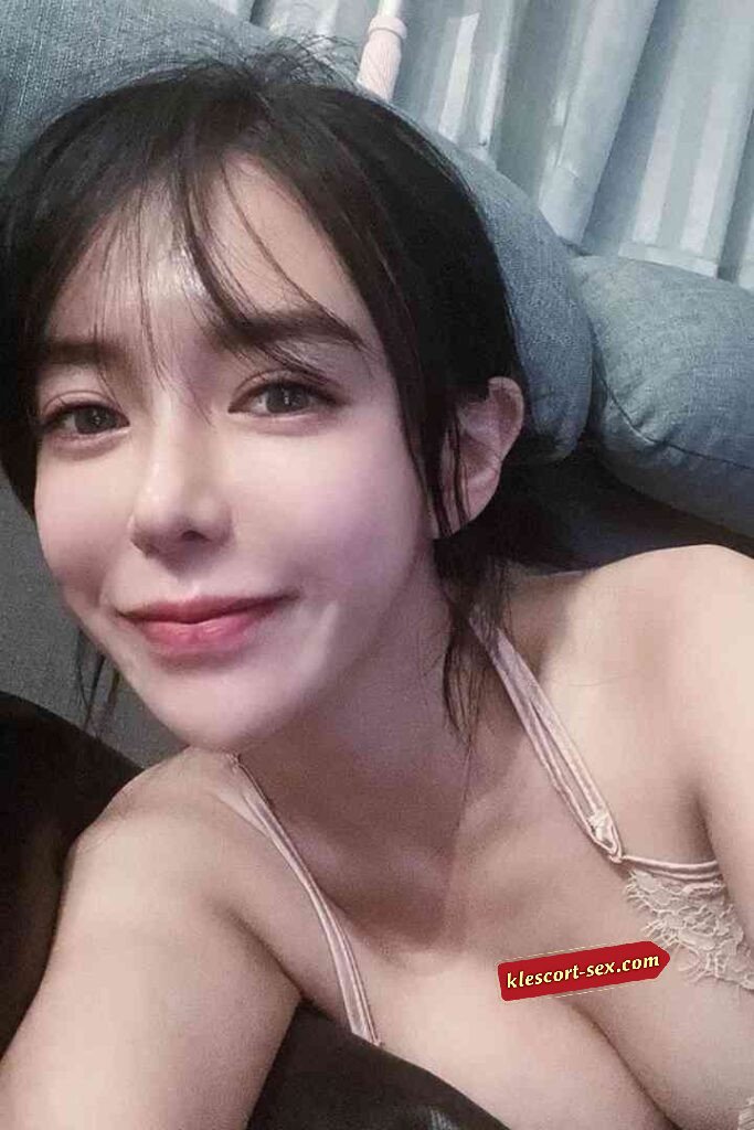 Smiling Asian woman with bangs, light makeup, and a lace top. Relaxed pose, soft lighting.