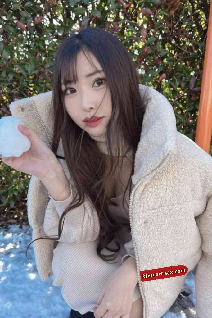 Asian woman in winter coat holds snowball outside with trees in background.