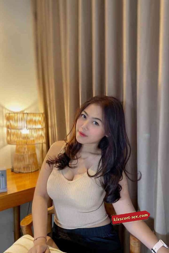 Attractive Asian woman in tank top sitting in room with lamp and curtains. Smiling at the camera.