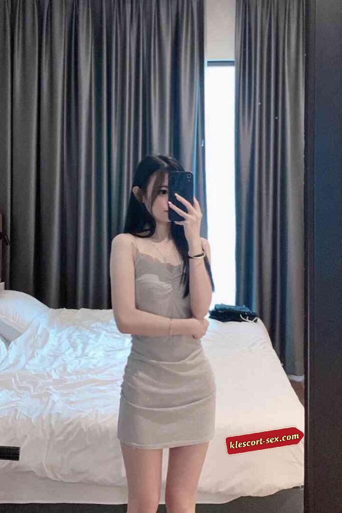 Woman in a beige dress takes a mirror selfie in a bedroom setting.