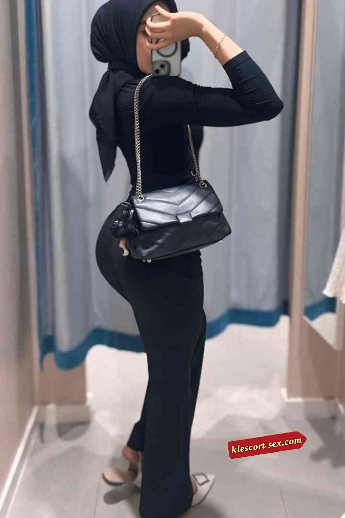 Woman in black dress and hijab taking mirror selfie with purse.