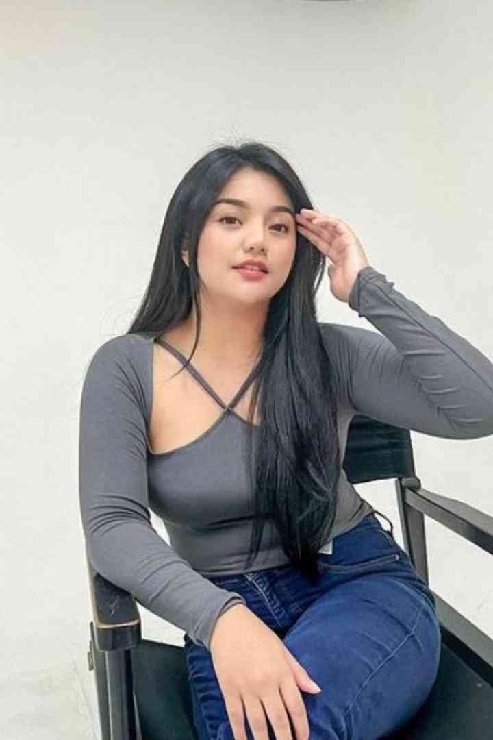 Beautiful woman with long black hair sitting in chair, wearing gray top and jeans, hand on head.