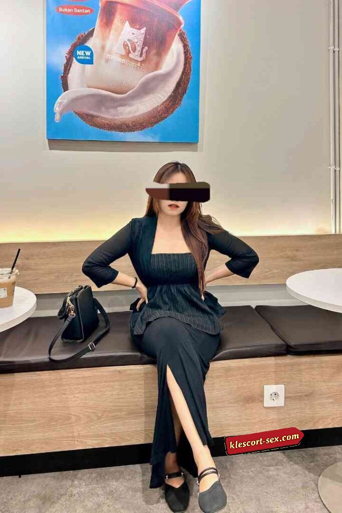 Woman in black outfit sitting on bench with a drink poster behind.