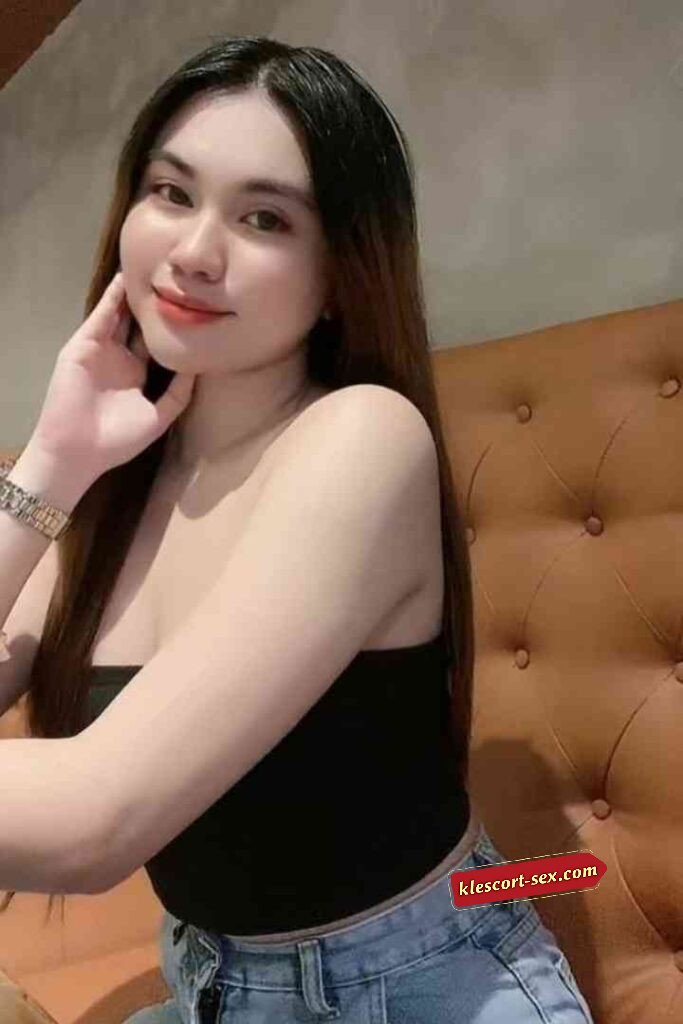 Smiling woman with long brown hair in black top and jeans posing on brown tufted couch.