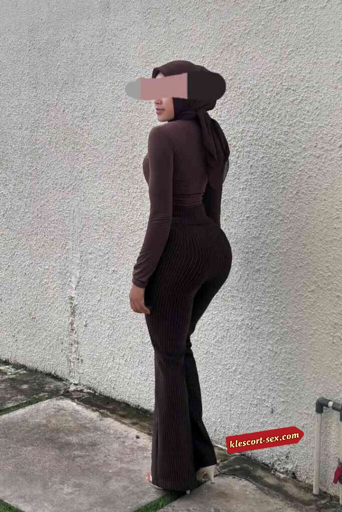 Woman in brown turtleneck and pants, wearing a hijab, standing outside.