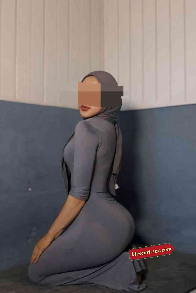 Woman in gray dress and hijab kneeling indoors, looking over shoulder.