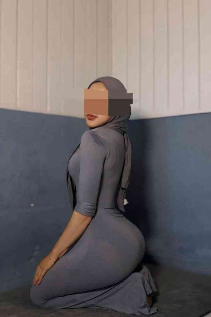 Woman kneeling, wearing grey hijab and dress, looking over shoulder.