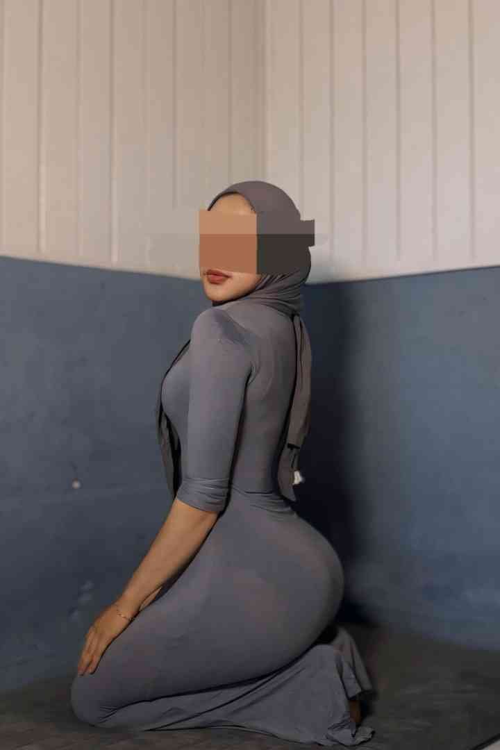 Woman kneeling, wearing grey hijab and dress, looking over shoulder.