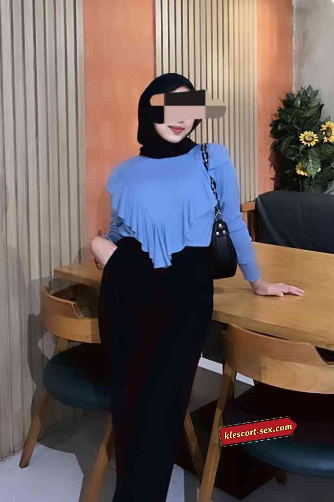 Woman in hijab, blue ruffled top, and black skirt posing indoors.