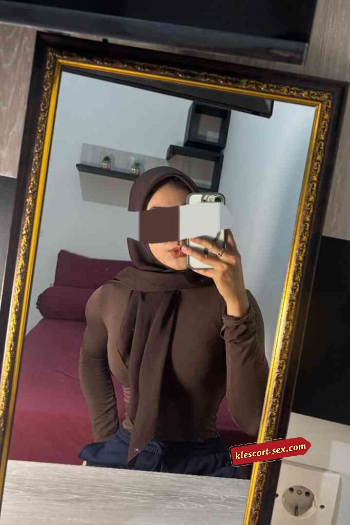 Woman in hijab takes mirror selfie in brown top. Interior background with bed and decor.