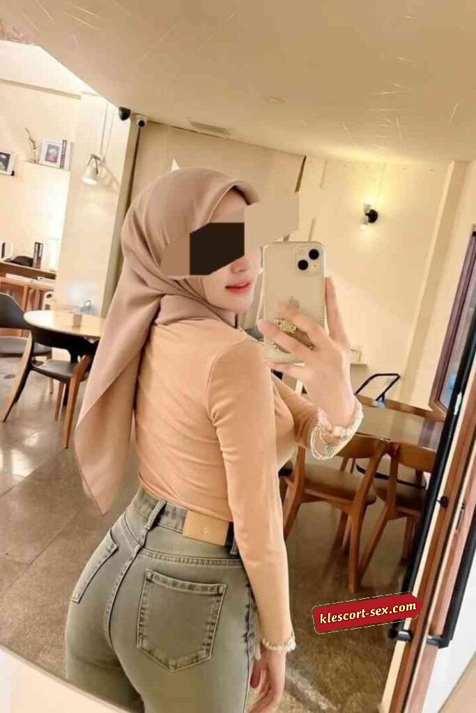 Woman in hijab taking mirror selfie, wearing jeans and a long-sleeved top.