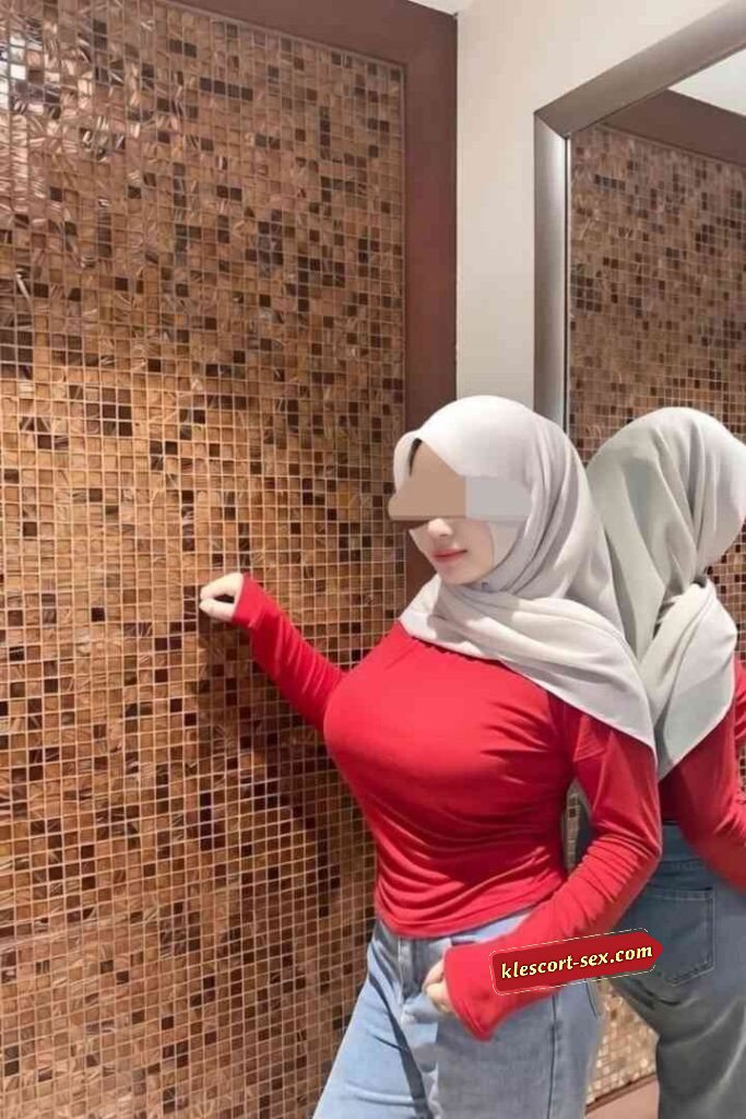 Woman in hijab, red shirt, and jeans posing by tiled wall.