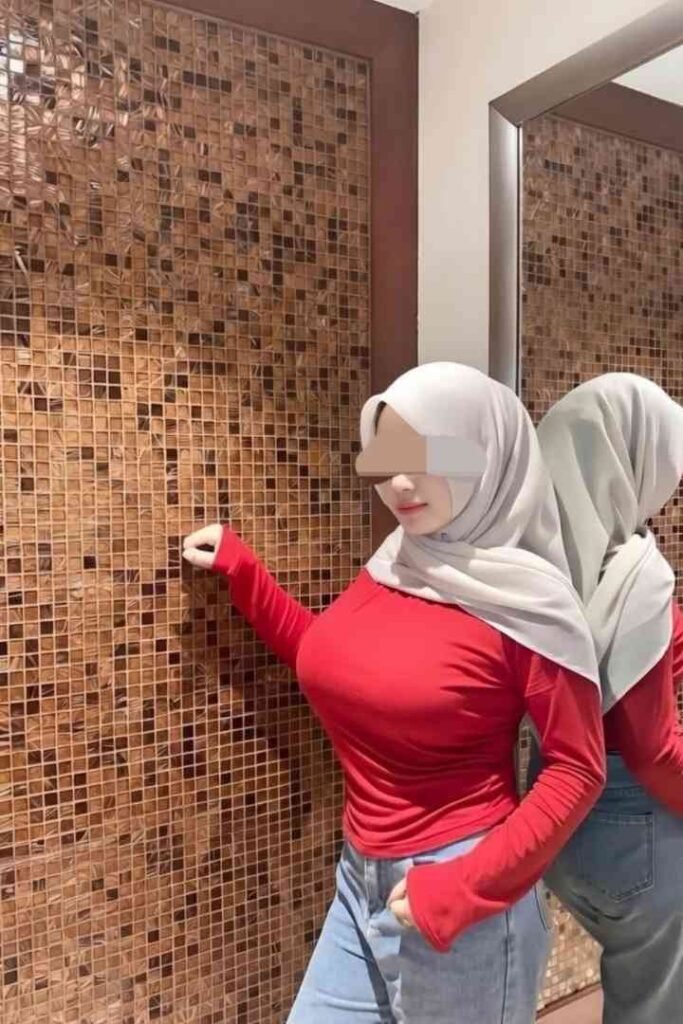 Woman in hijab, red shirt, and jeans posing by tiled wall with mirror reflection.