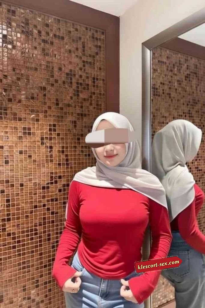 Woman in hijab, red top, and jeans posing in front of a mirror.