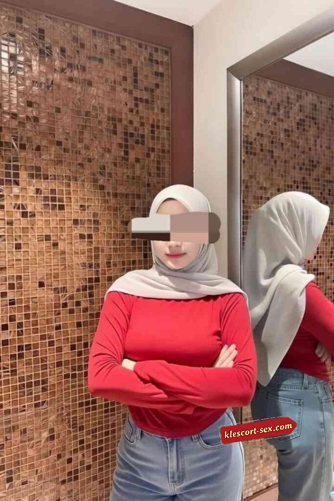 Woman in hijab, red top, and jeans posing in front of mirror.