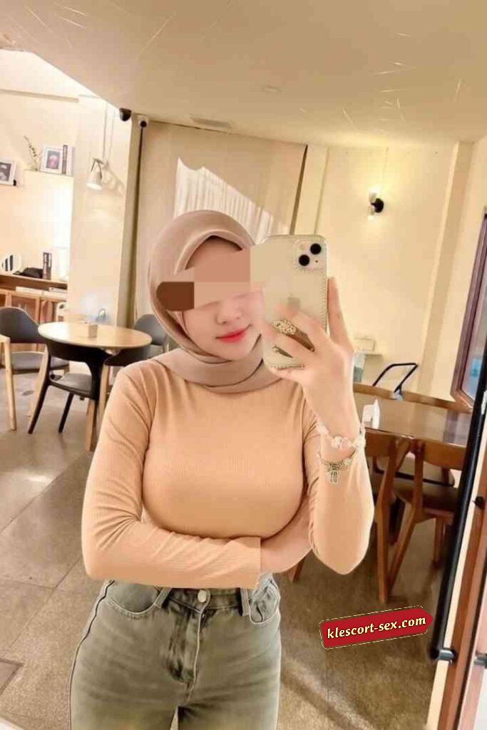 Woman in hijab taking selfie indoors. Beige top, jeans, cafe setting.