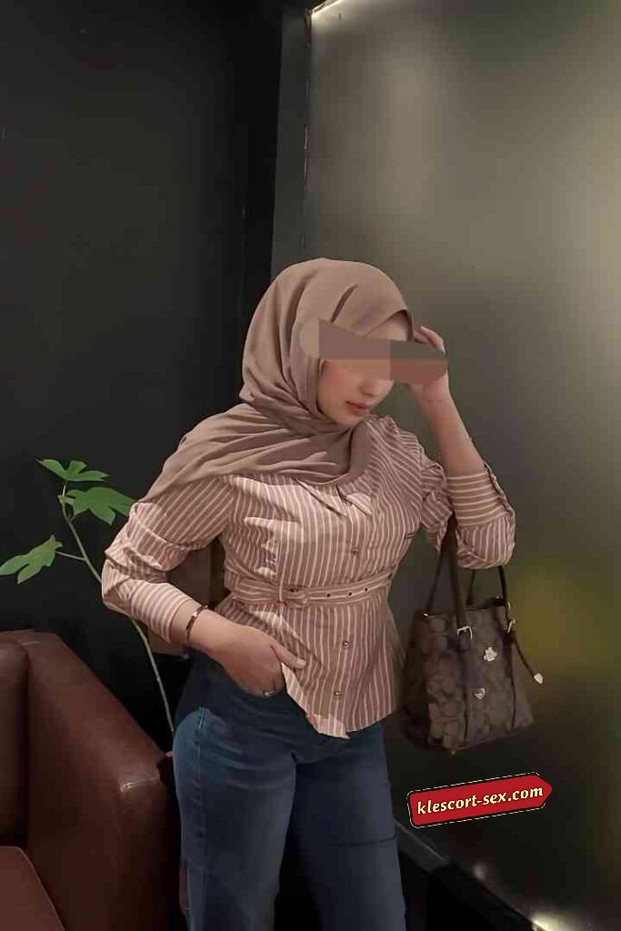 Woman in hijab, striped shirt, jeans, and handbag stands indoors.