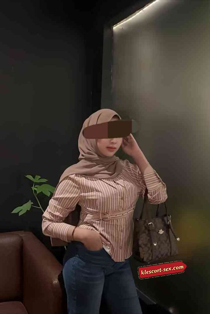 Woman in hijab with striped shirt, jeans, and purse standing indoors.