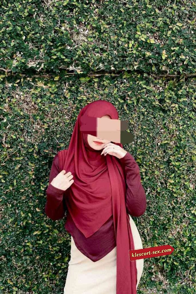 Woman in maroon hijab and long-sleeved shirt poses against green wall.