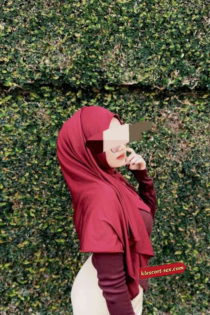 Woman in maroon hijab against green foliage wall, posing with hand near face.