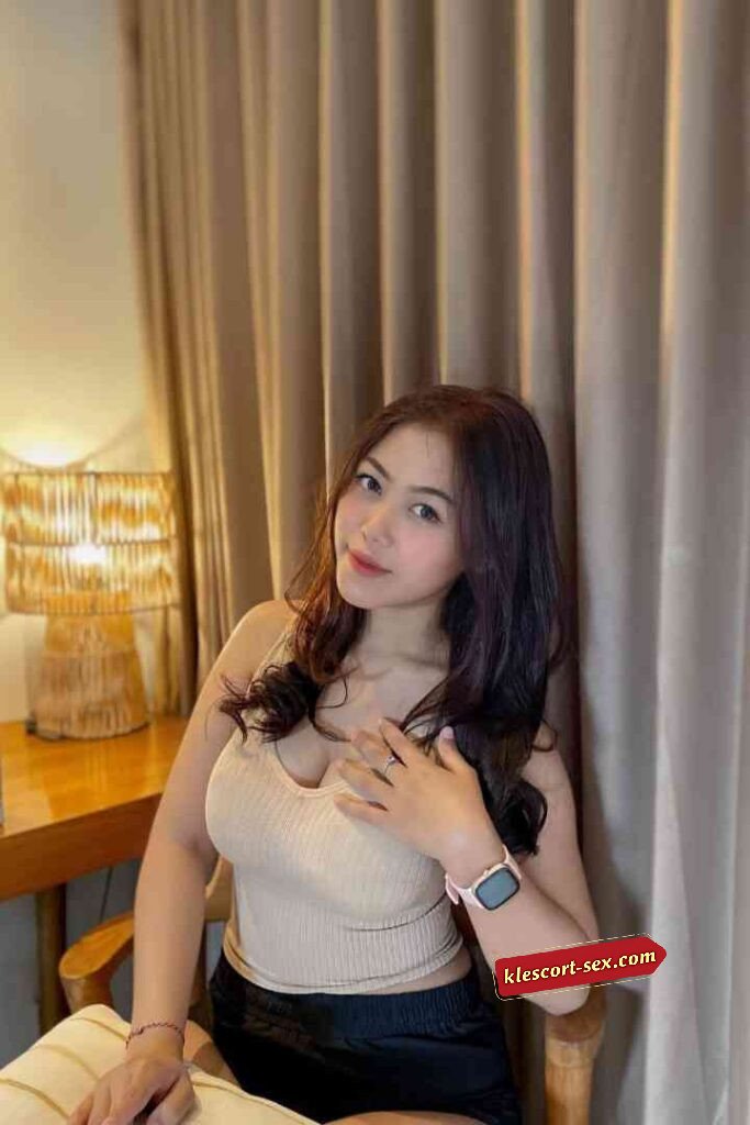 Attractive woman in a tank top and shorts poses indoors with soft lighting and neutral tones.