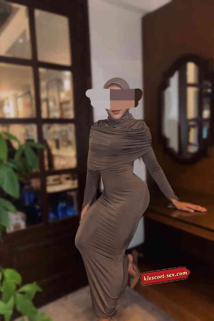 Woman in draped taupe modest dress and hijab posing indoors. Elegant, long-sleeved gown.