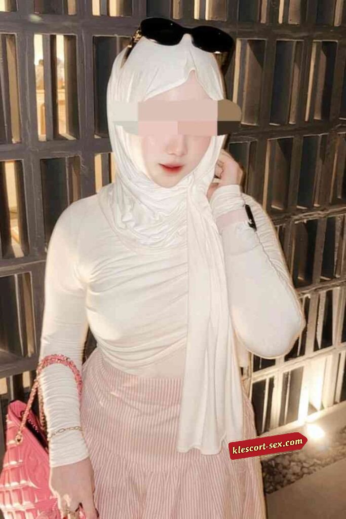 Woman in white hijab, pink skirt, holding pink purse.