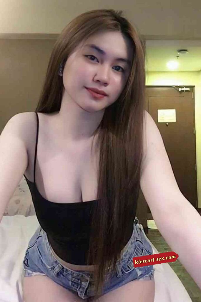 Attractive young woman with long brown hair in a black top and denim shorts posing indoors.