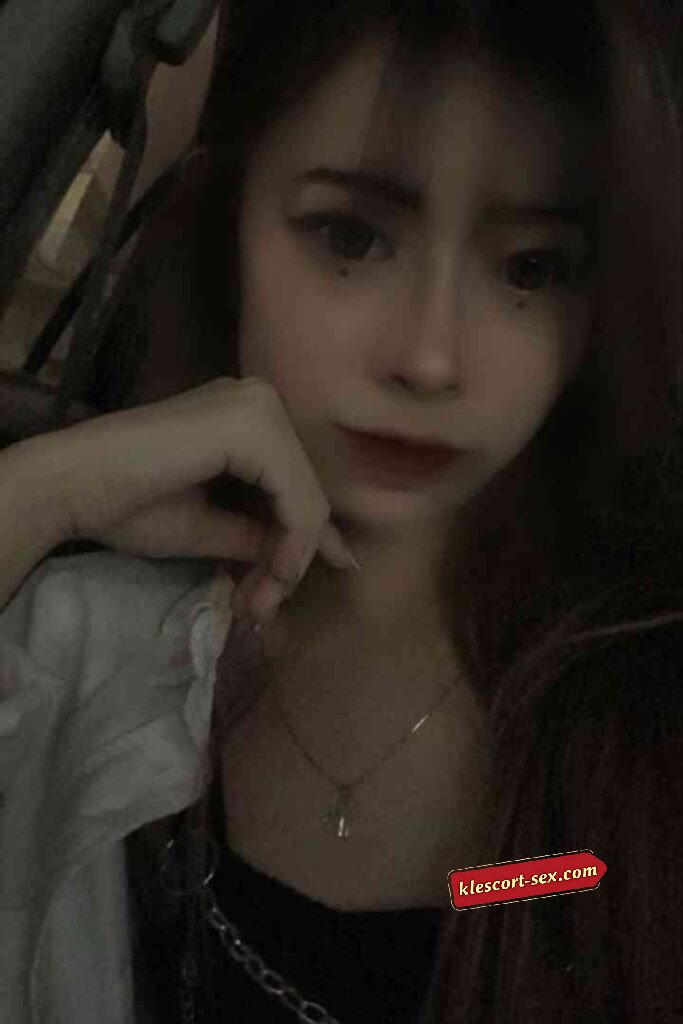 Young woman with dark hair, dark lipstick, and a delicate necklace posing indoors.