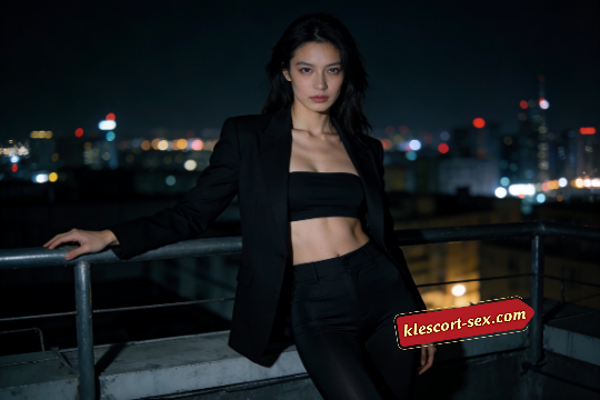 Woman in black suit on rooftop at night with city lights in background.