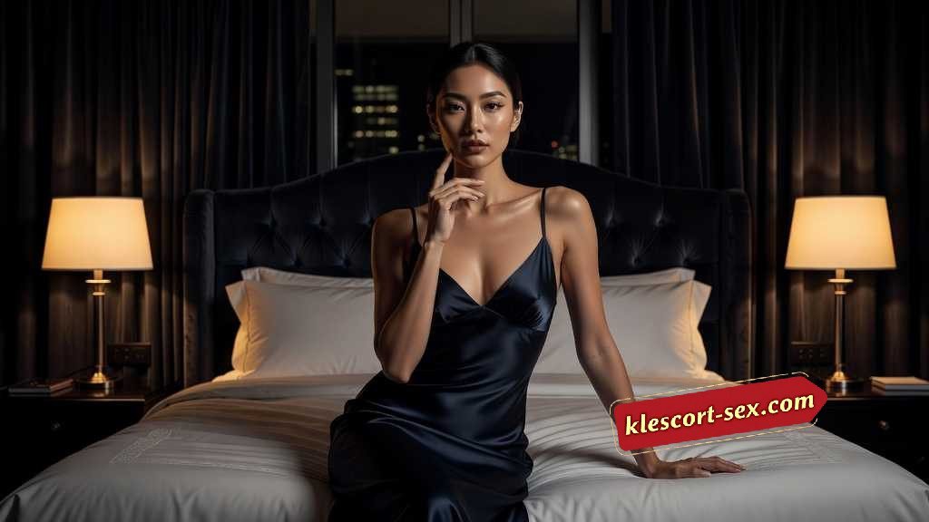 KL Escort Girl Elegant woman in a black slip dress sits on a bed in a dimly lit room. Sex Service Malaysia