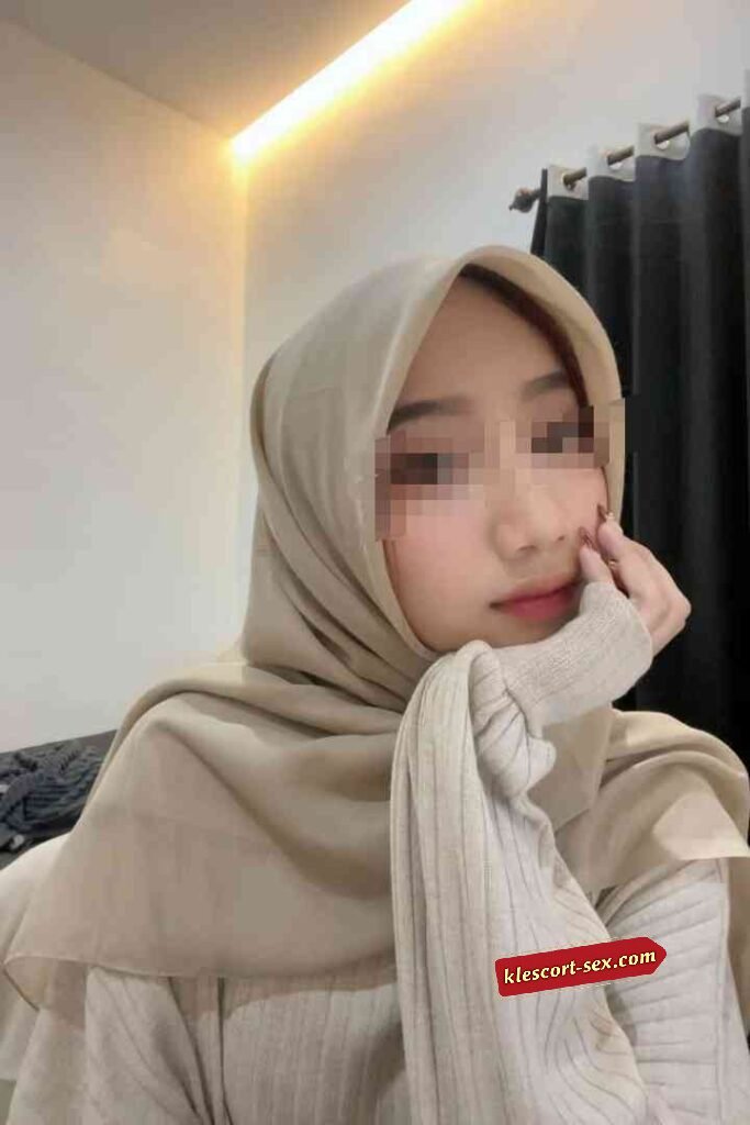 KL Escort Girl Young woman in a beige hijab and sweater, resting her chin on her hand. Sex Service Malaysia
